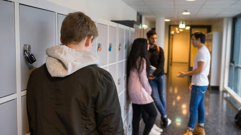 Tenåring i grå hettegenser og svart jakke står lent inntil ei rekk med nøkkelskap, med hodet ned. Litt bortafor i gangen står ei gruppe med andre elever.